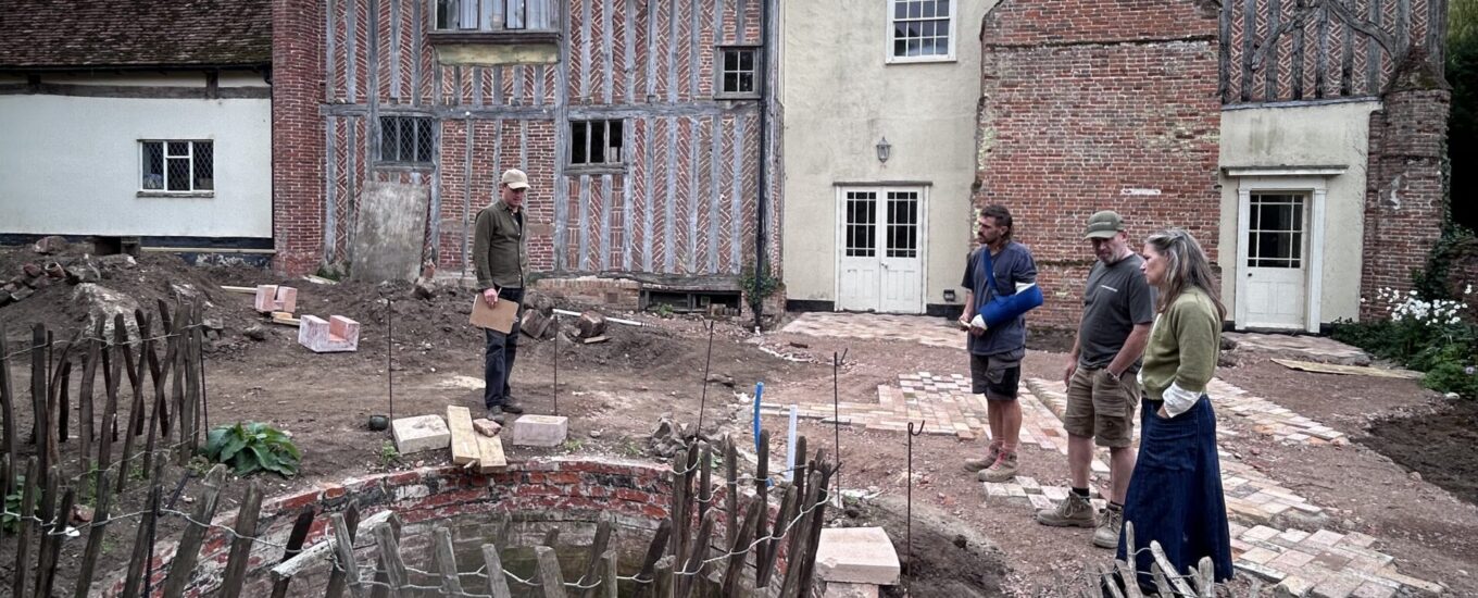 An image with historic house, Benton End, in the background under a grey sky. Four people stand around the pond-to-be in the midground.