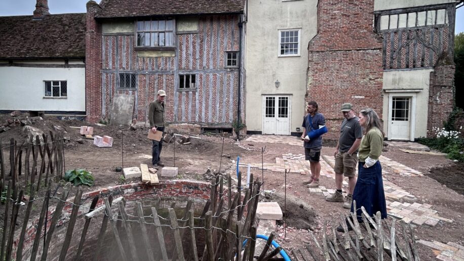 An image with historic house, Benton End, in the background under a grey sky. Four people stand around the pond-to-be in the midground.
